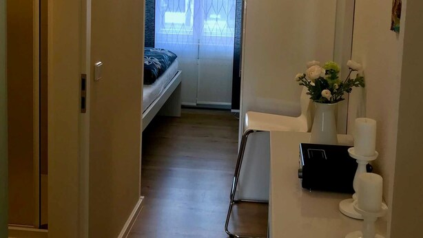 A narrow hallway with a bed in the background, a white desk with a vase of flowers, and a window with a view of buildings.