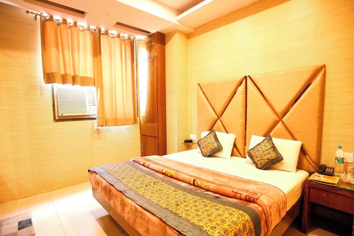 A hotel room with a large bed, a wooden headboard, a bedside table with a lamp, and a window with curtains.