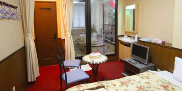 Une chambre d’hôtel comprenant un lit, une télévision, une petite table avec une chaise, une fenêtre avec des rideaux et une salle de bain avec un lavabo et un miroir.