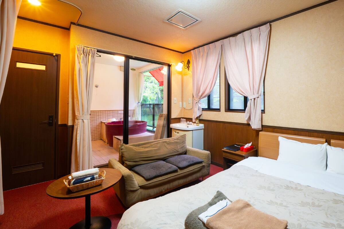 A hotel room with a bed, a sofa, a small table, a nightstand, a window with curtains, and a view of a red jacuzzi outside.