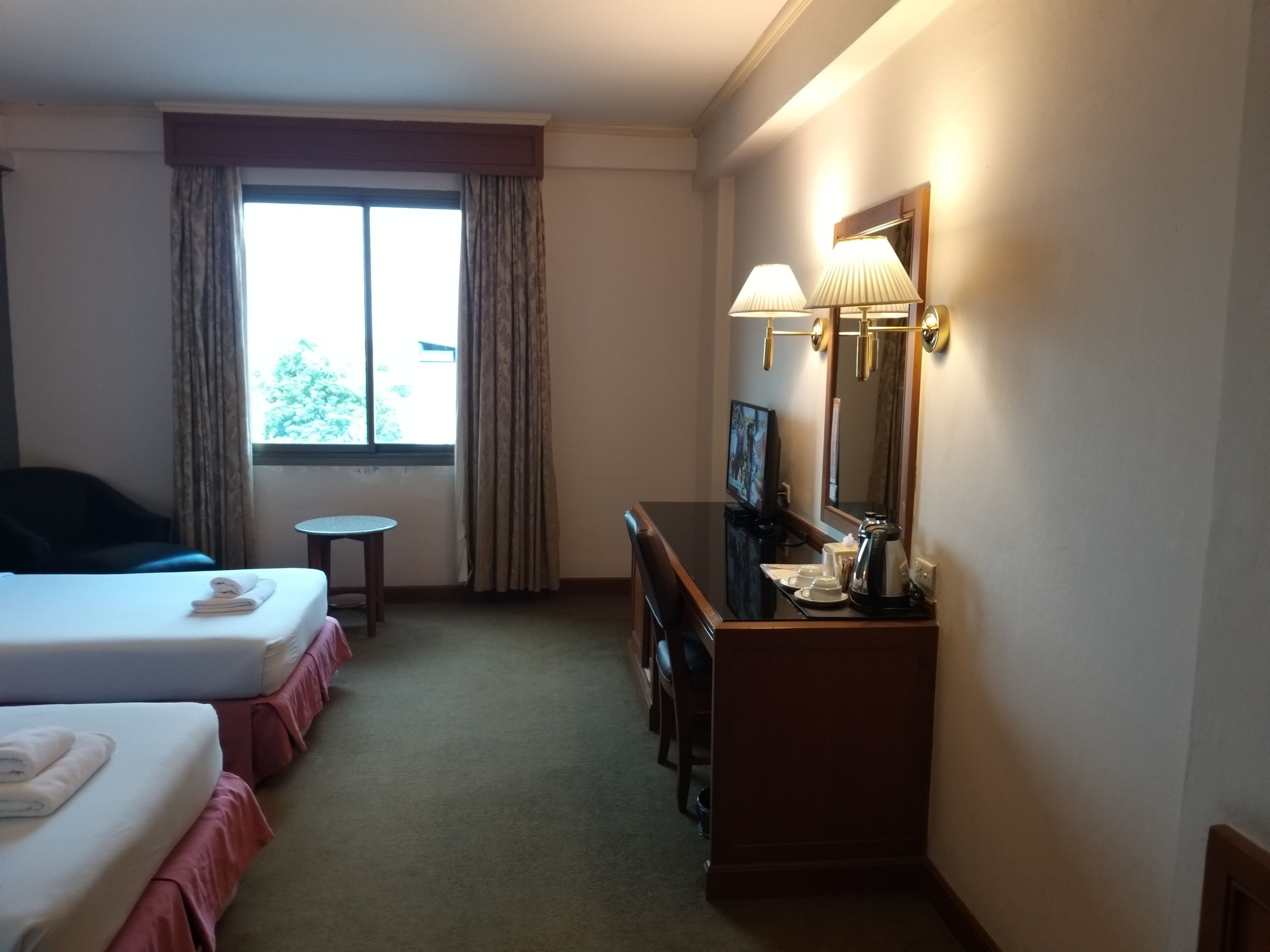 Ein Hotelzimmer mit zwei Betten, einem Schreibtisch mit Spiegel, einem Fernseher und einem Fenster mit Vorhängen.