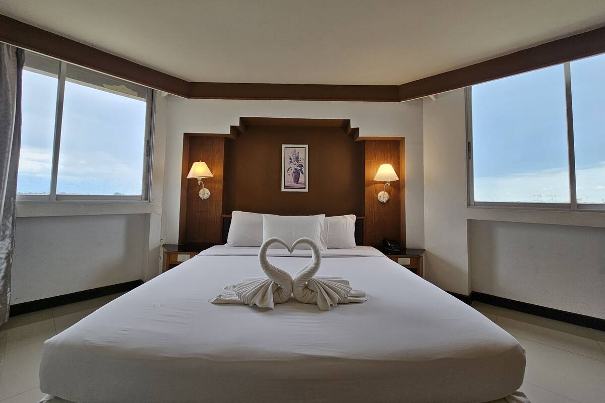 A hotel room with a large bed, two bedside lamps, a framed picture on the wall, and a view of the sea through the windows.