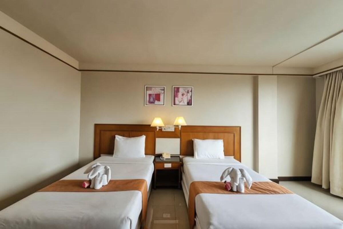 A hotel room with two single beds, a wooden headboard, bedside lamps, a nightstand, and a window with curtains.
