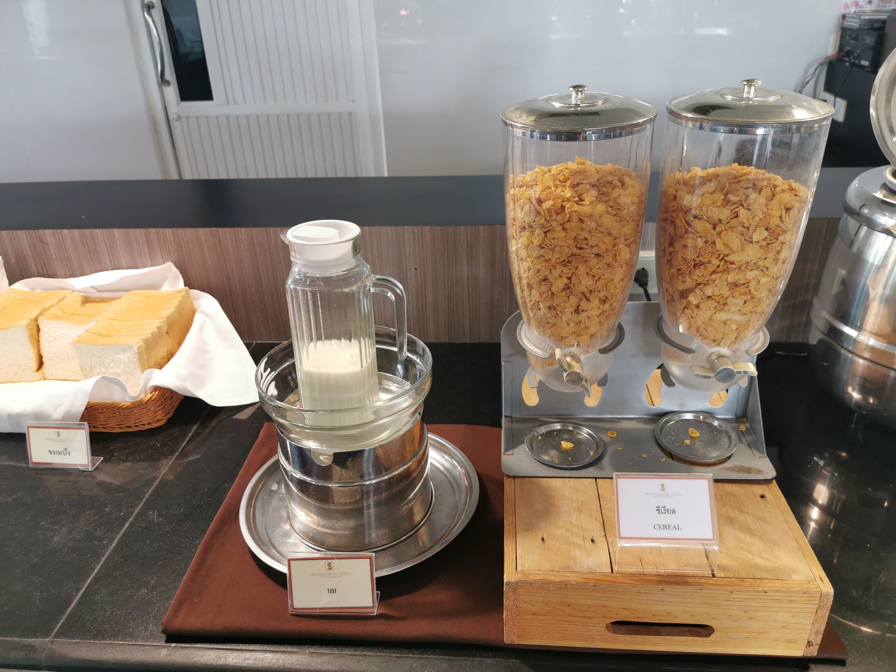 A buffet setup with a milk dispenser, cereal dispensers, and a bread basket.