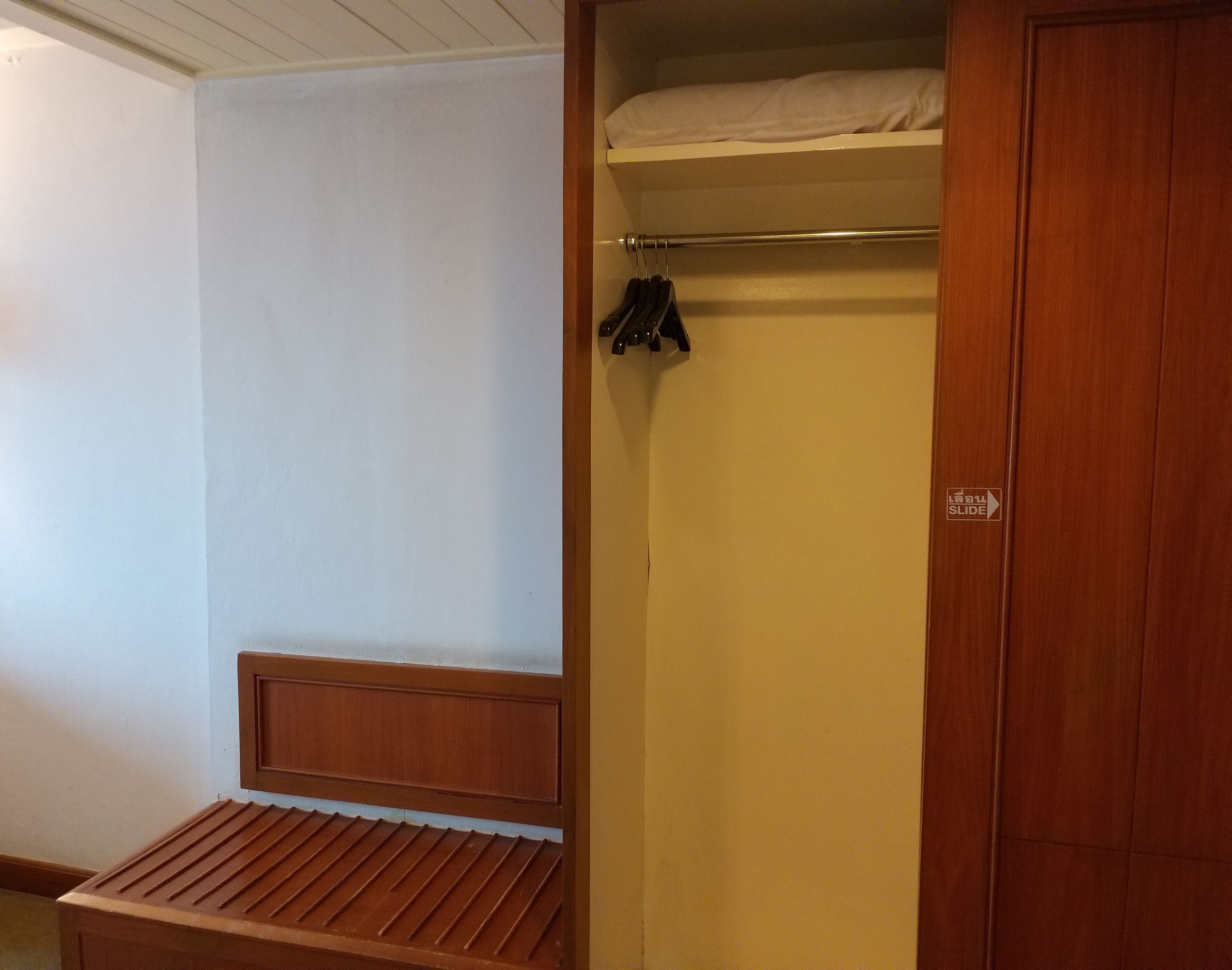Een kamer met een houten bankje, een kledingkast met een schuifdeur en een bed met een wit laken.