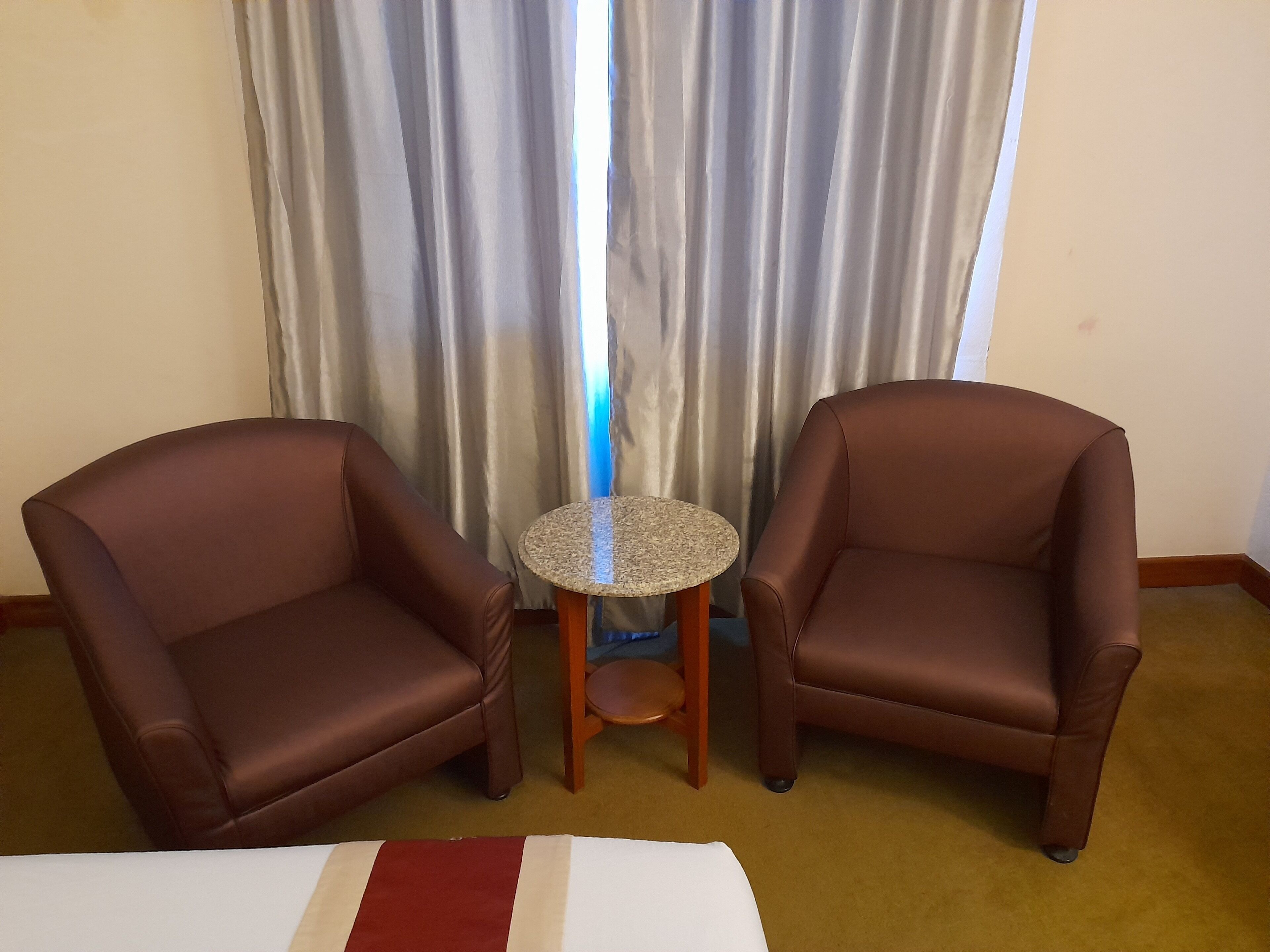 Dos sillones de cuero marrón enfrentados, con una mesita redonda entre ellos, una cama blanca con franja roja, y una ventana con cortinas grises.