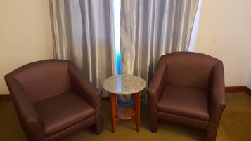 Dos sillones de cuero marrón enfrentados, con una mesita redonda entre ellos, una cama blanca con franja roja, y una ventana con cortinas grises.