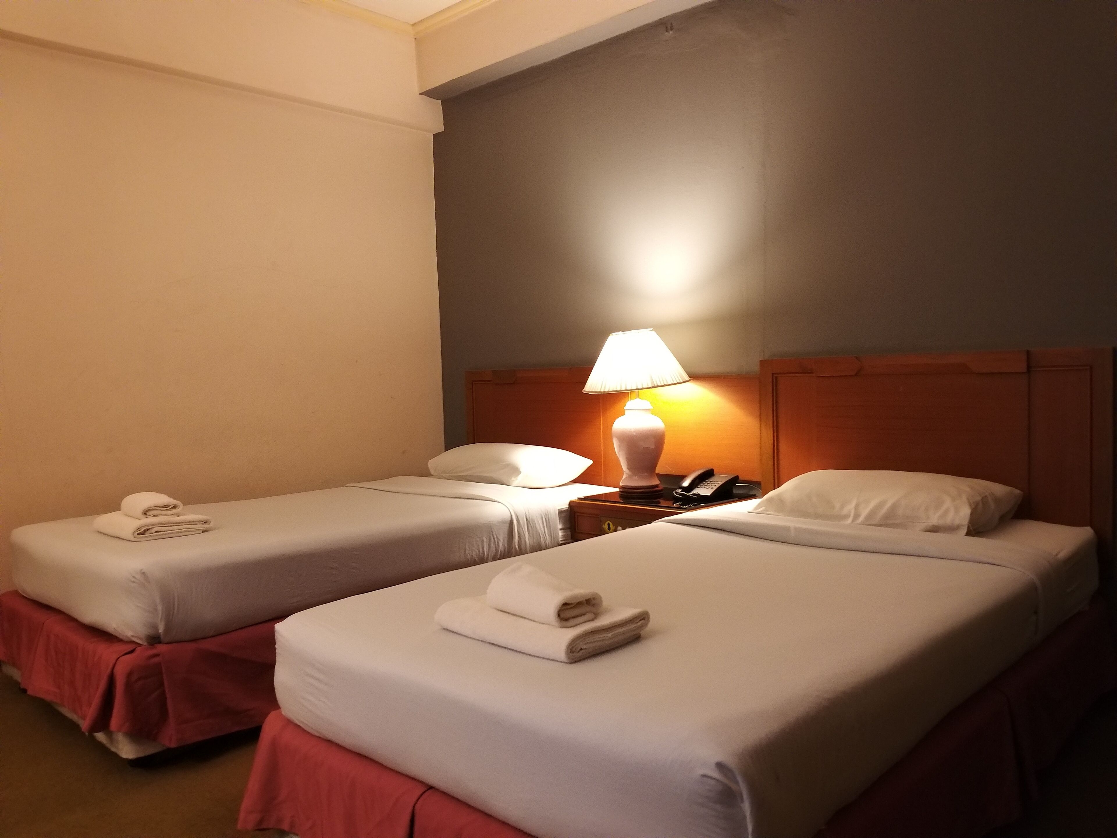 Habitación de hotel con dos camas, cada una con una almohada y una toalla doblada. Se observa una mesita de noche con una lámpara y un teléfono.