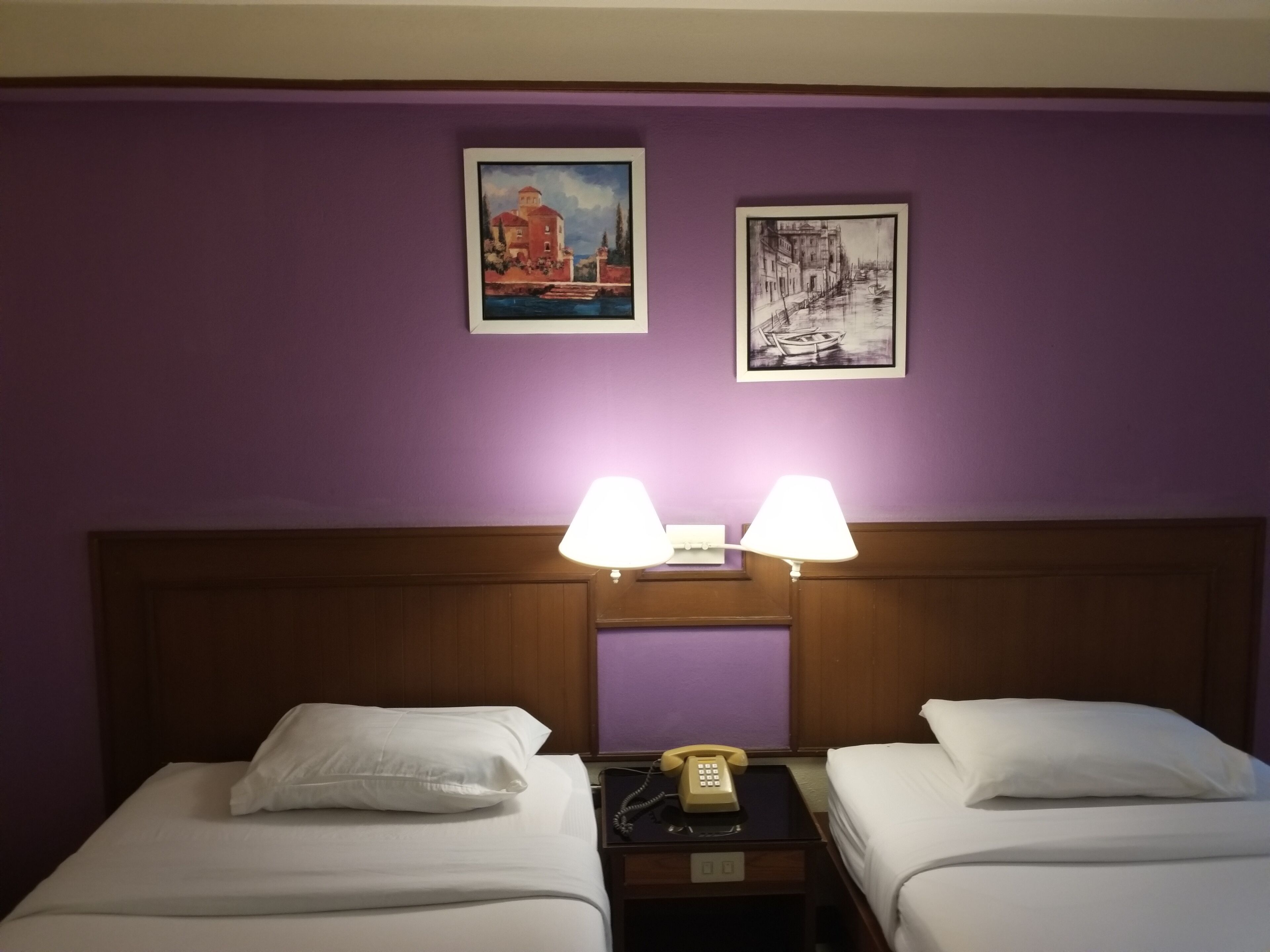 A hotel room with two beds, a wooden headboard, two wall-mounted lamps, and two framed pictures on the wall.