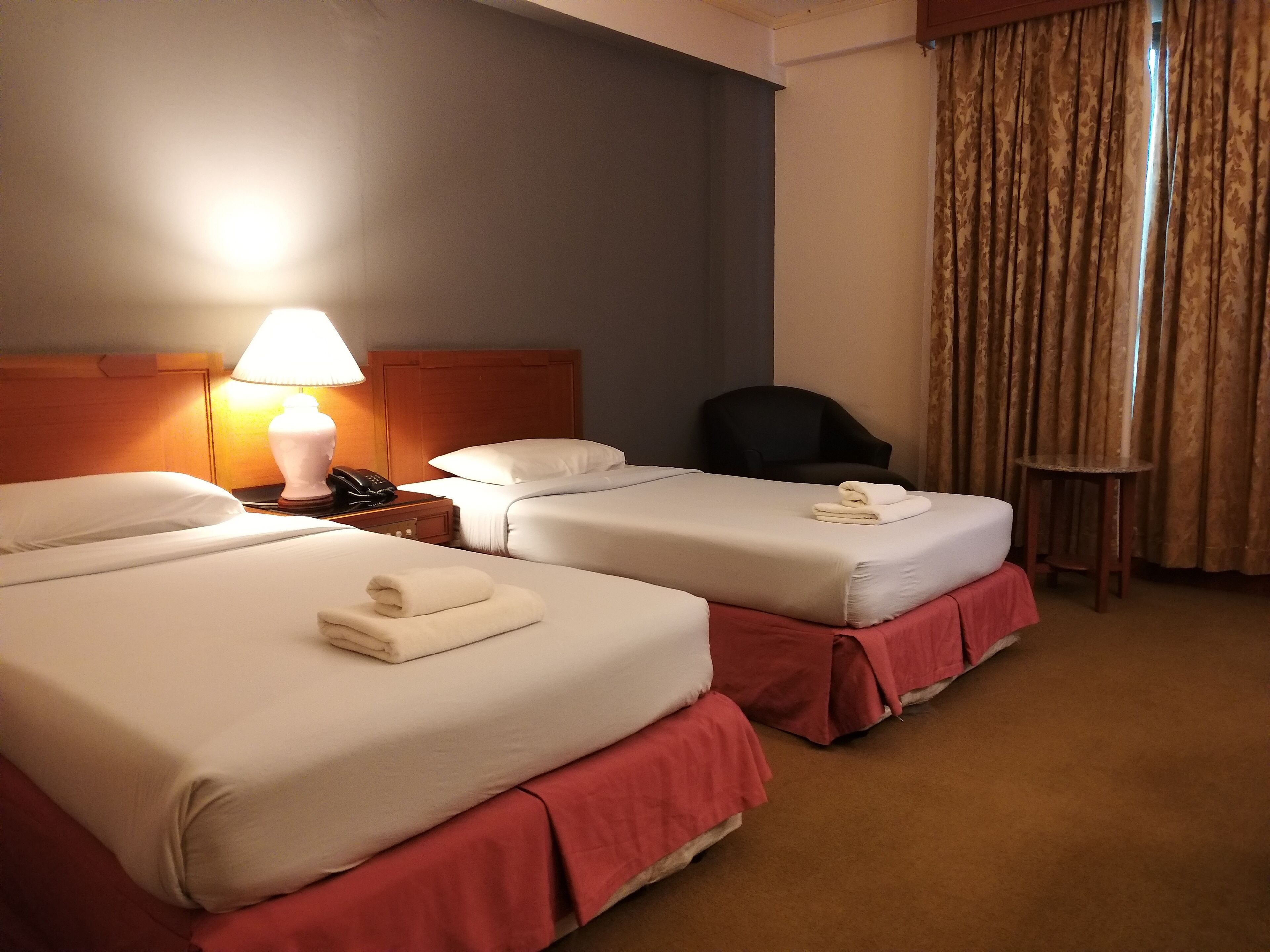 A hotel room with two beds, a nightstand with a lamp, a chair, and a small table.
