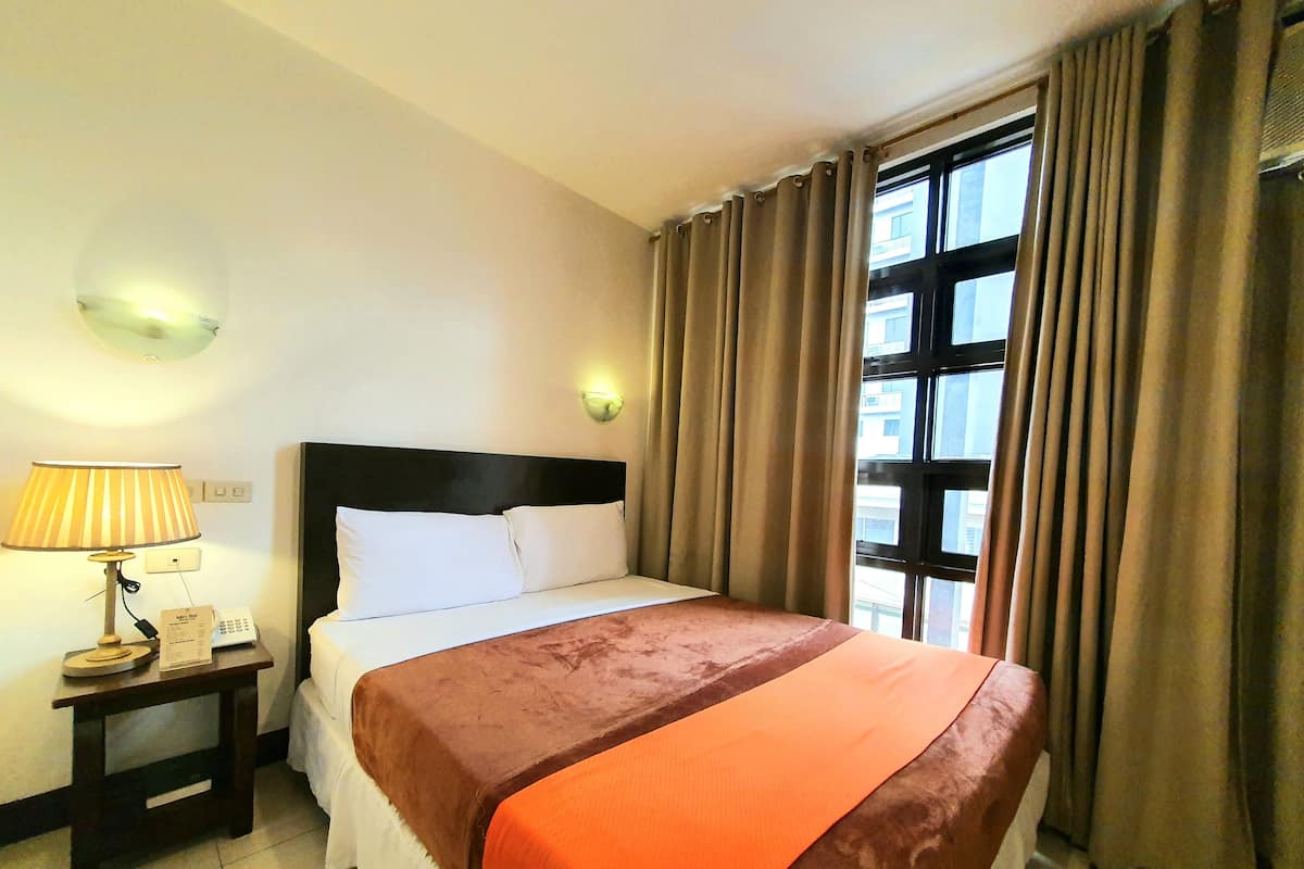 Habitación de hotel con cama, mesita de noche, lámpara, luz en la pared y ventana con cortinas.