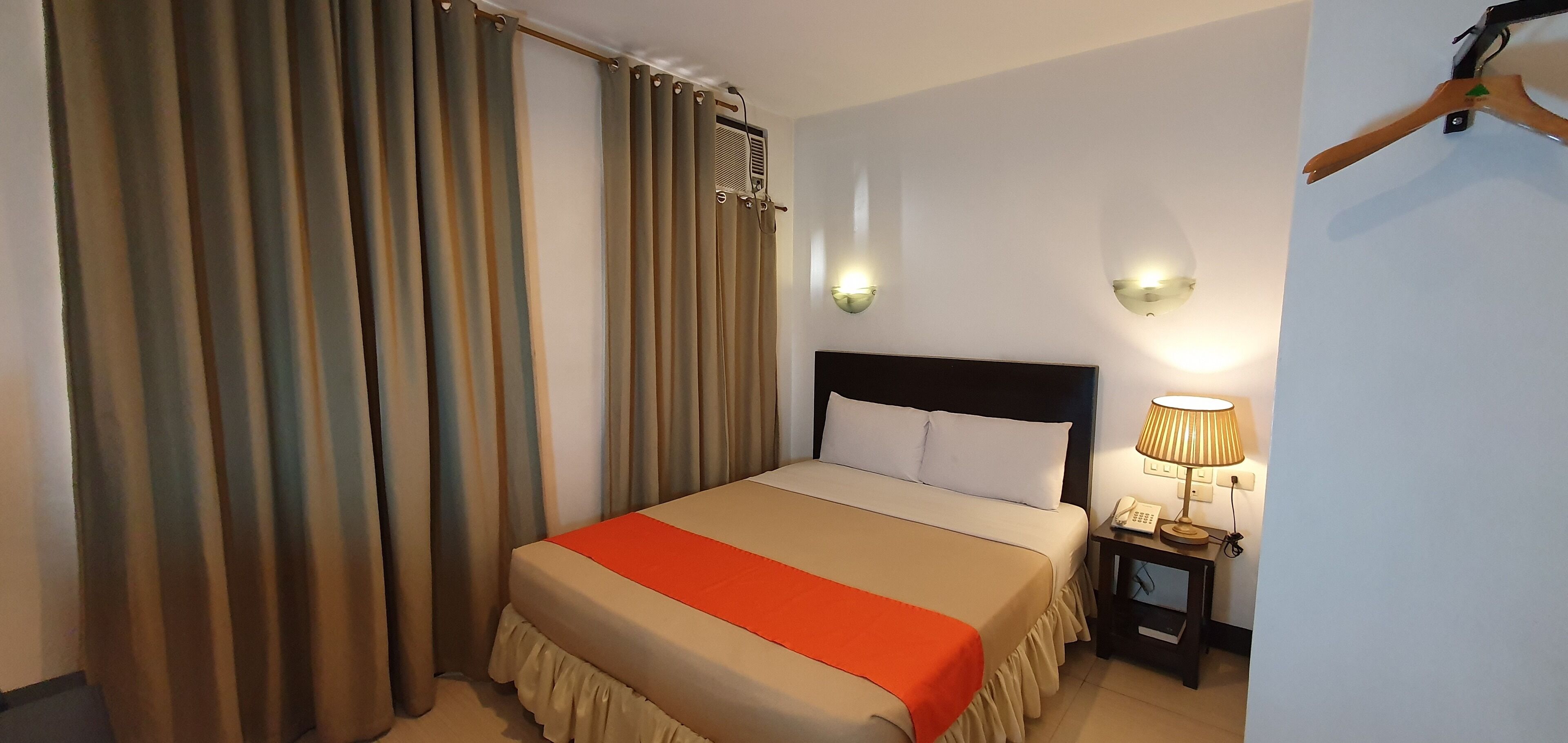 A hotel room with a bed, curtains, a nightstand with a lamp, and a wall-mounted light fixture.