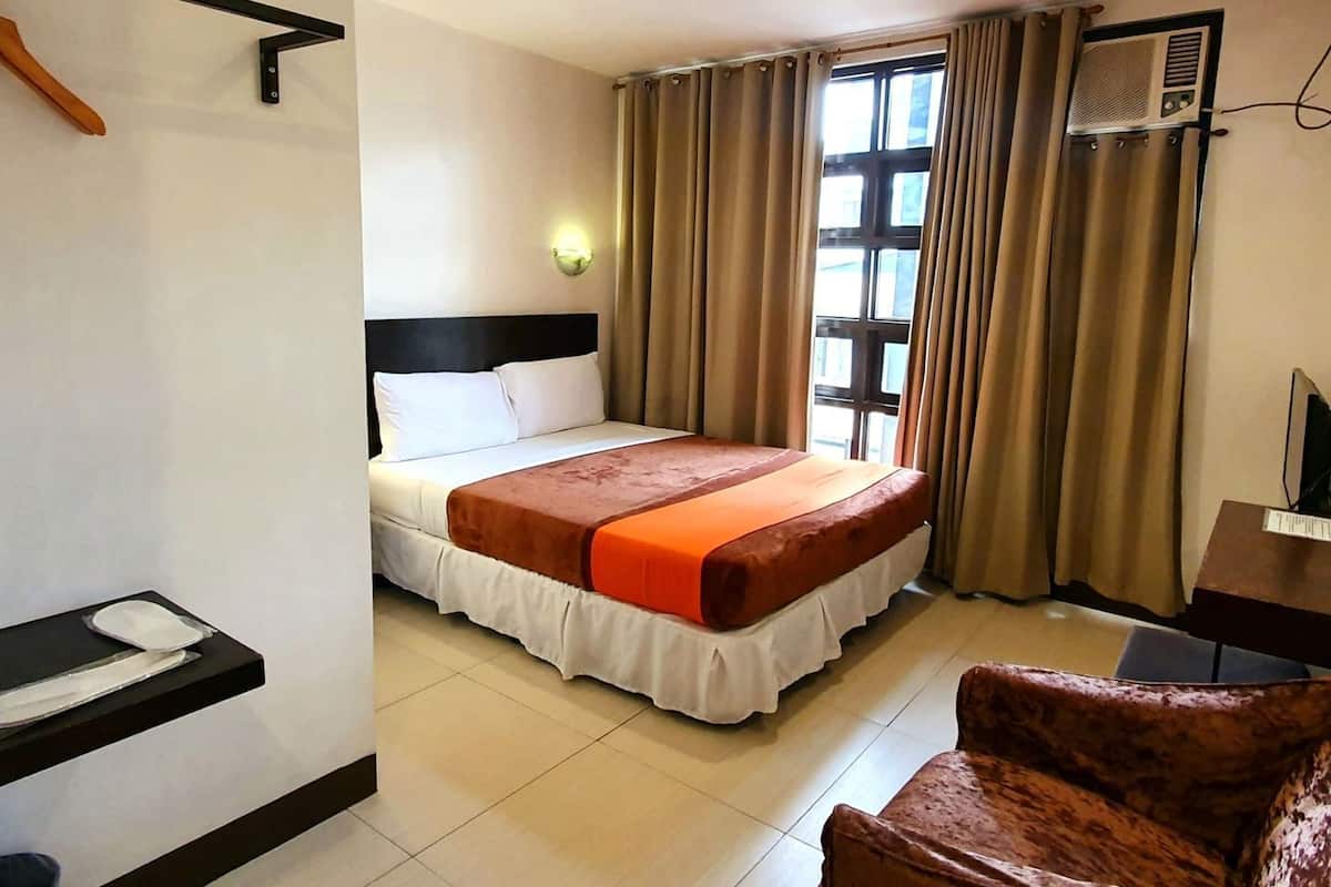 Una habitación de hotel con cama, silla, escritorio, ventana con cortinas y aire acondicionado.