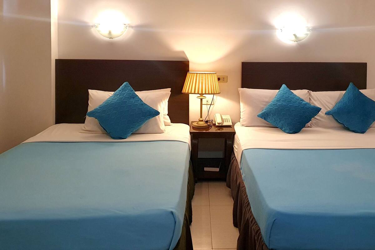 Habitación de hotel con dos camas individuales, cada una con colcha azul y almohadas a juego. Entre las camas hay una mesita de noche con una lámpara y un teléfono.