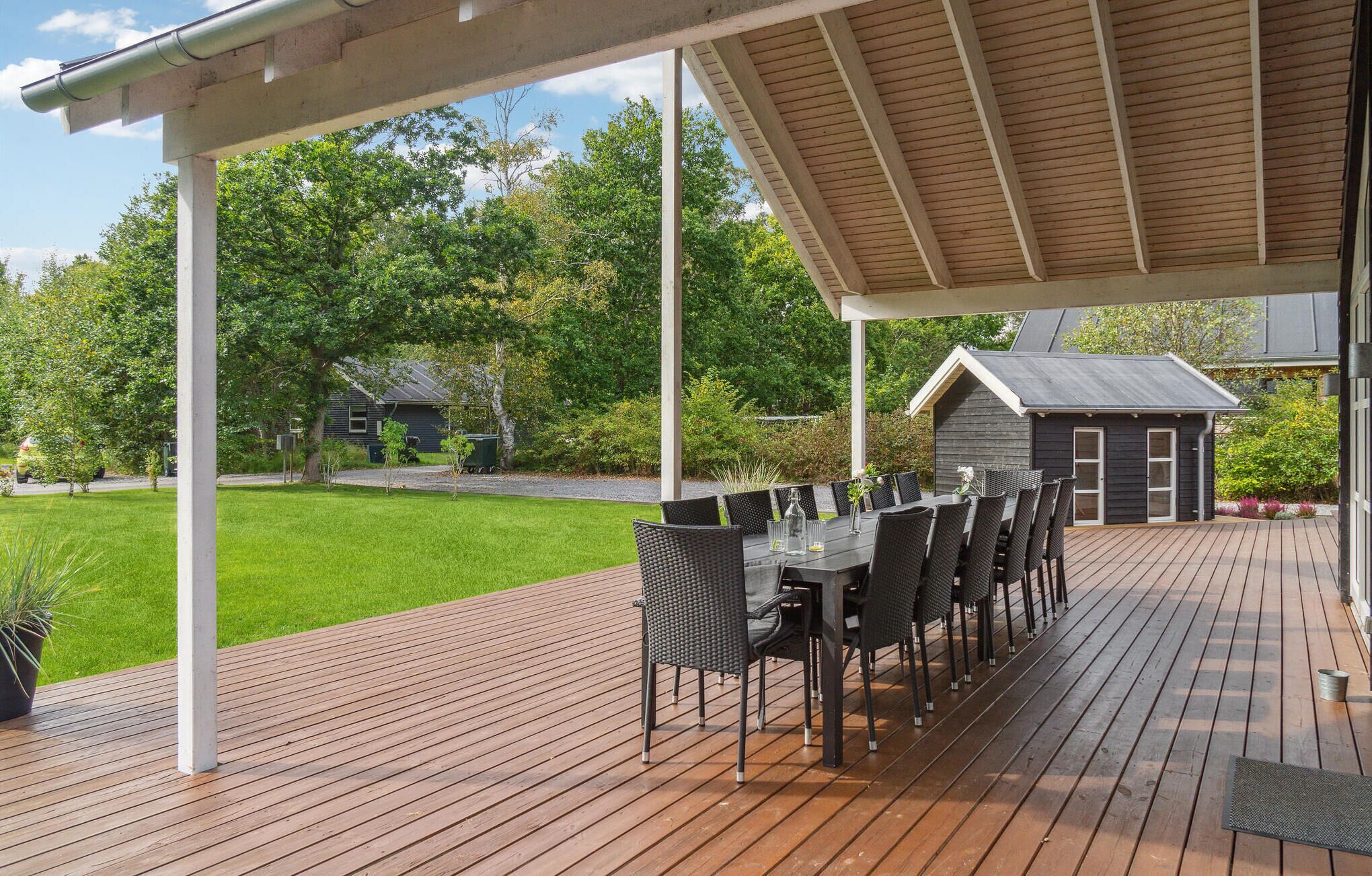 Een overdekt terras buiten met een eettafel en stoelen, omringd door een houten terras en een gazon.
