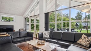 A spacious living room with a large black leather sofa, a wooden coffee table, and a TV on the left. Large windows provide a view of the outdoors. - Gorgeous home in Grenaa with sauna (Grenaa)
