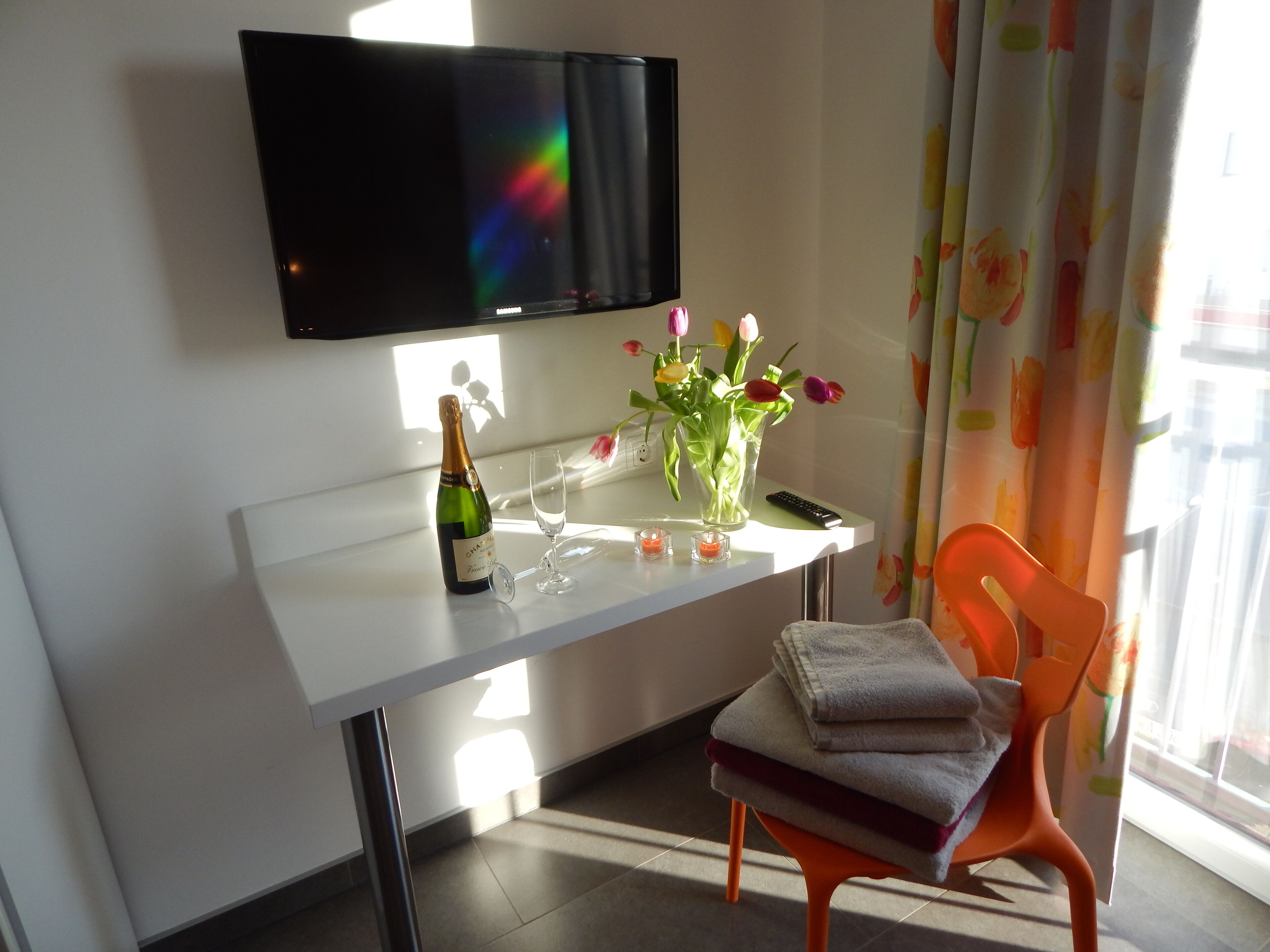 Une chambre d’hôtel avec un bureau blanc, un téléviseur fixé au mur, un vase avec des fleurs, une bouteille de champagne, un verre et une télécommande.