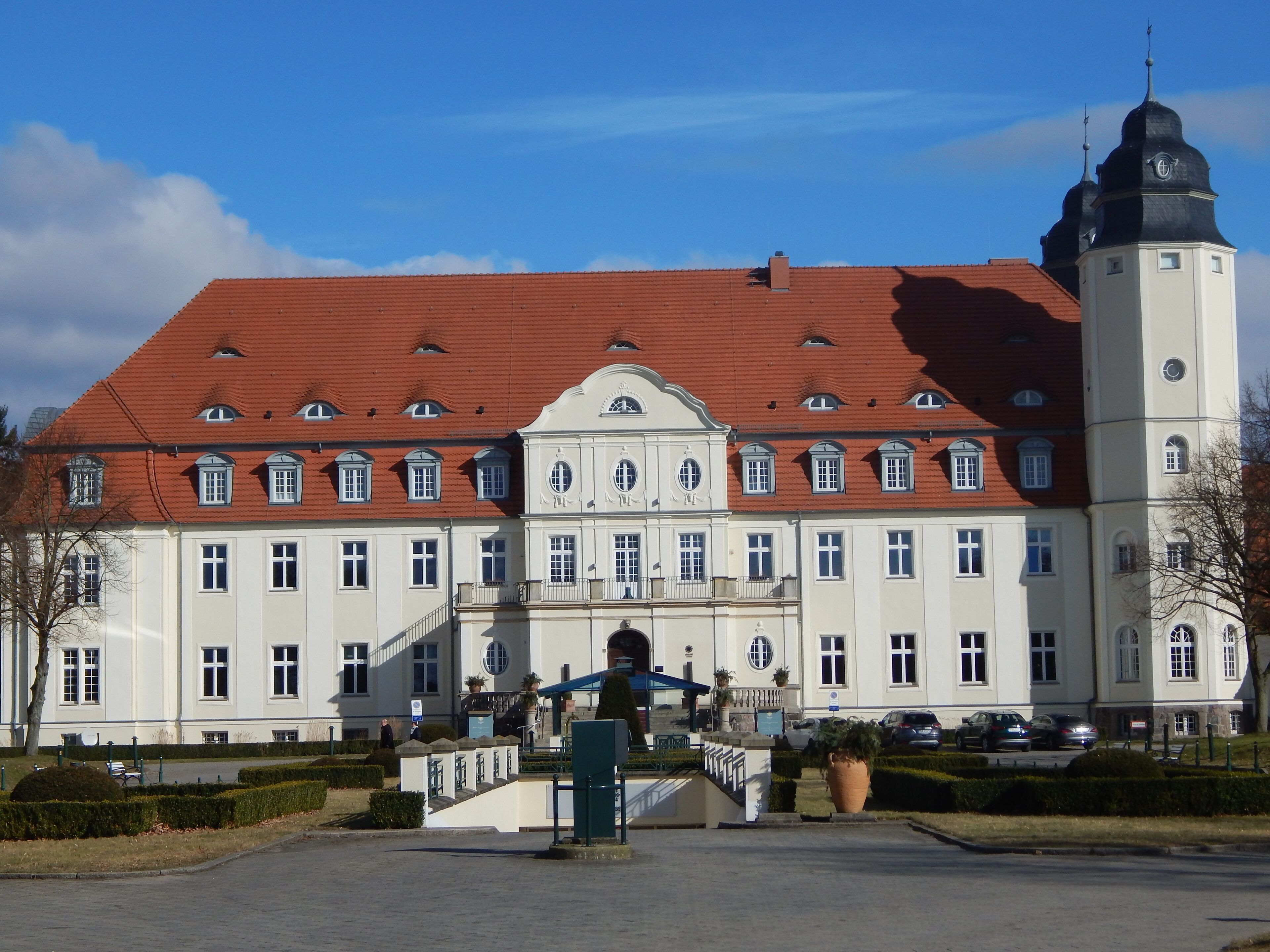 A large, historic building with a red-tiled roof, multiple windows, and a prominent tower with a clock.