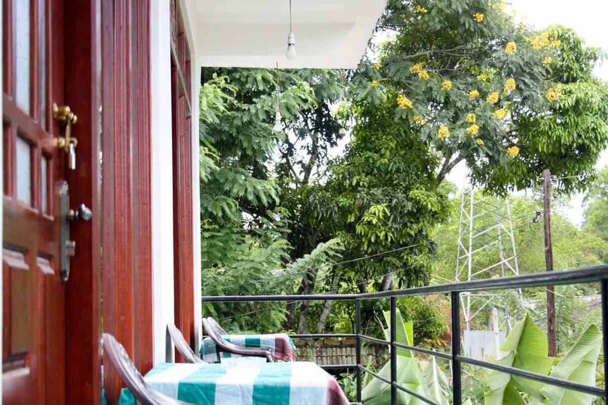 A balcony with a checkered tablecloth, a metal railing, and a view of lush greenery and yellow flowers.