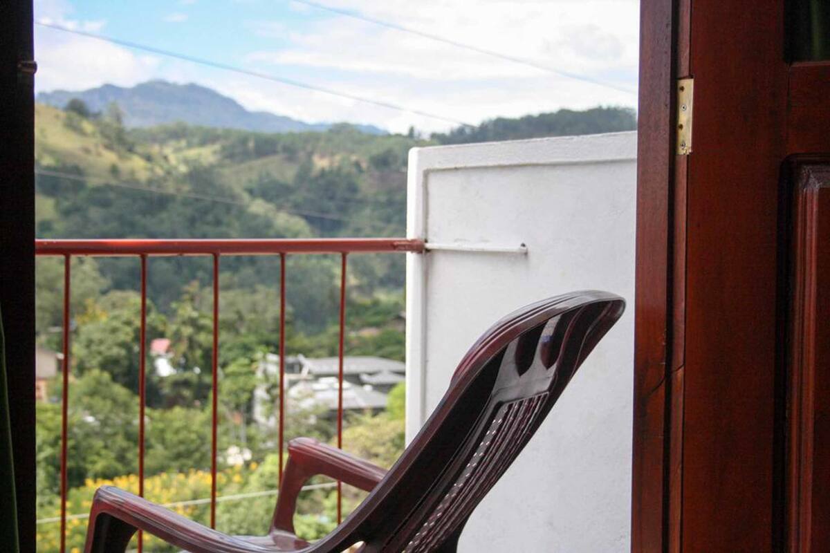 A balcony with a red railing, a white wall, and a view of green hills and buildings in the background.