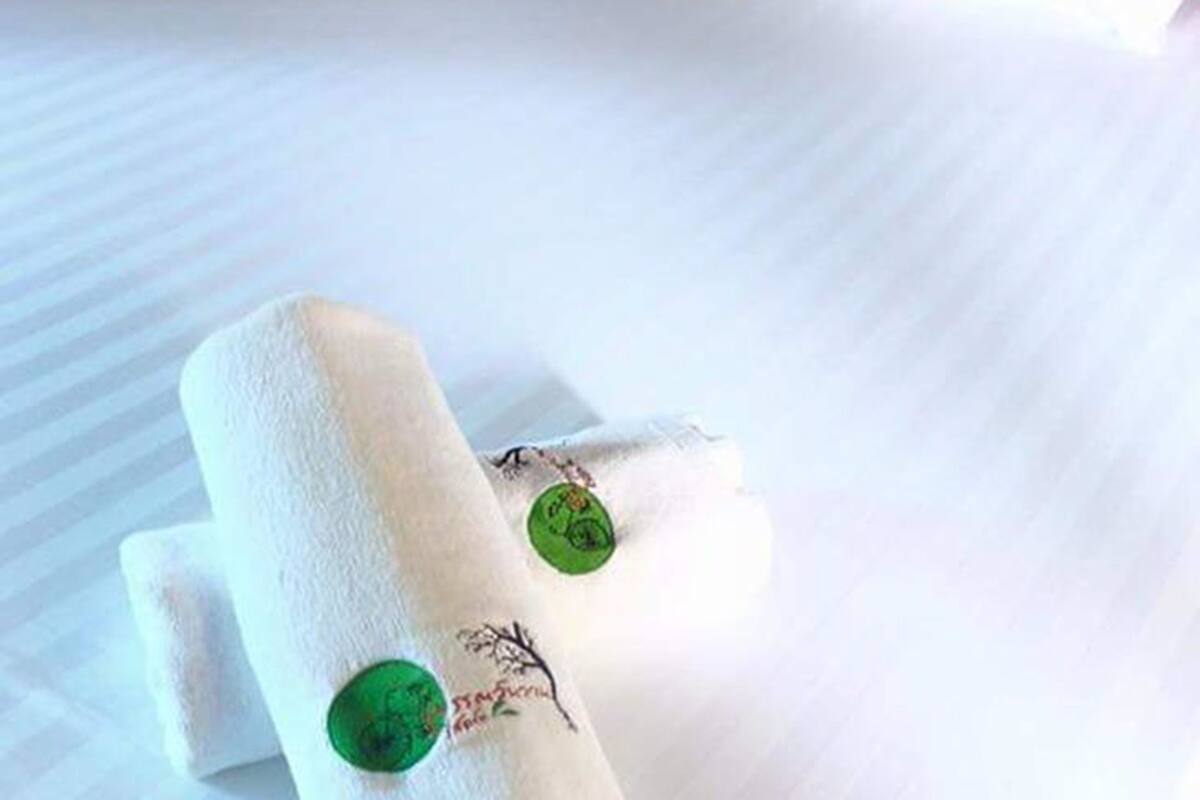 Un lit bien fait, avec des draps blancs, et une serviette enroulée, ornée d’un bouton vert et d’un petit motif brodé.