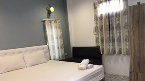 A neatly made bed with white linens, a headboard, and a rolled towel on the bed. There is a wooden wardrobe to the right and a wall-mounted light fixture above the bed.