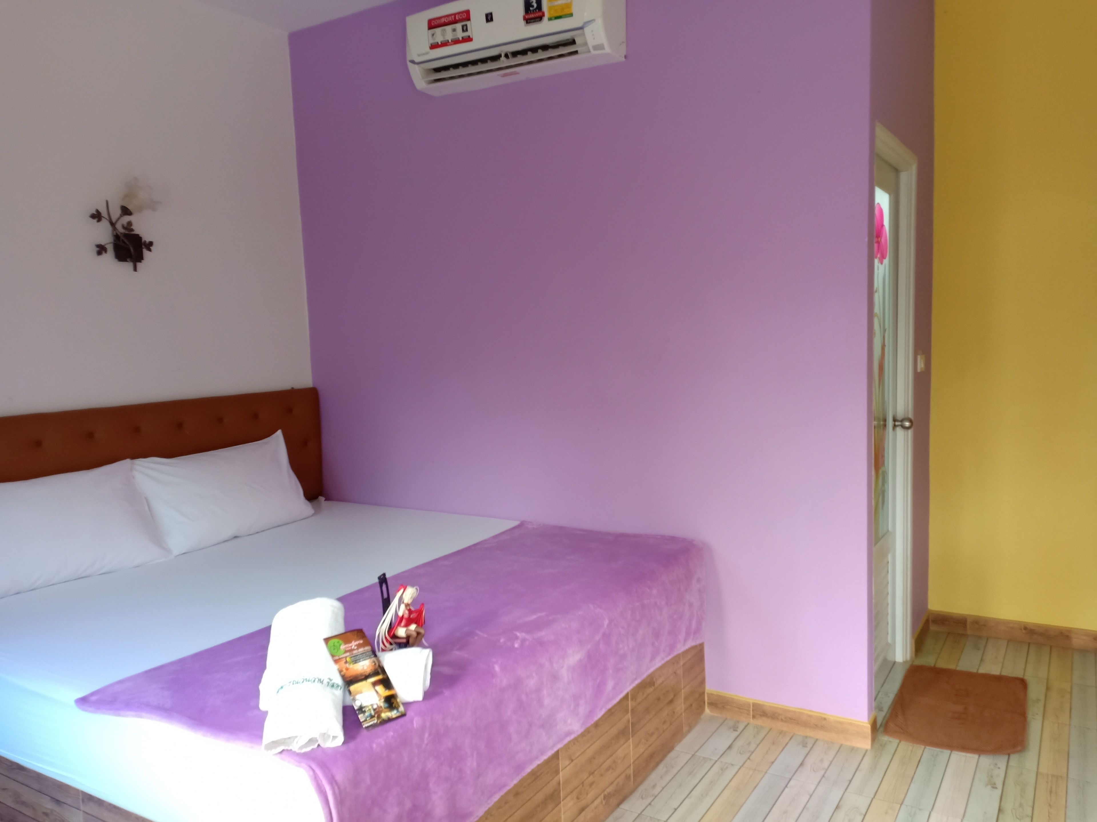 A hotel room with a bed, a purple wall, a wooden headboard, a white pillow, a white towel, a small statue, a magazine, and a brown mat.