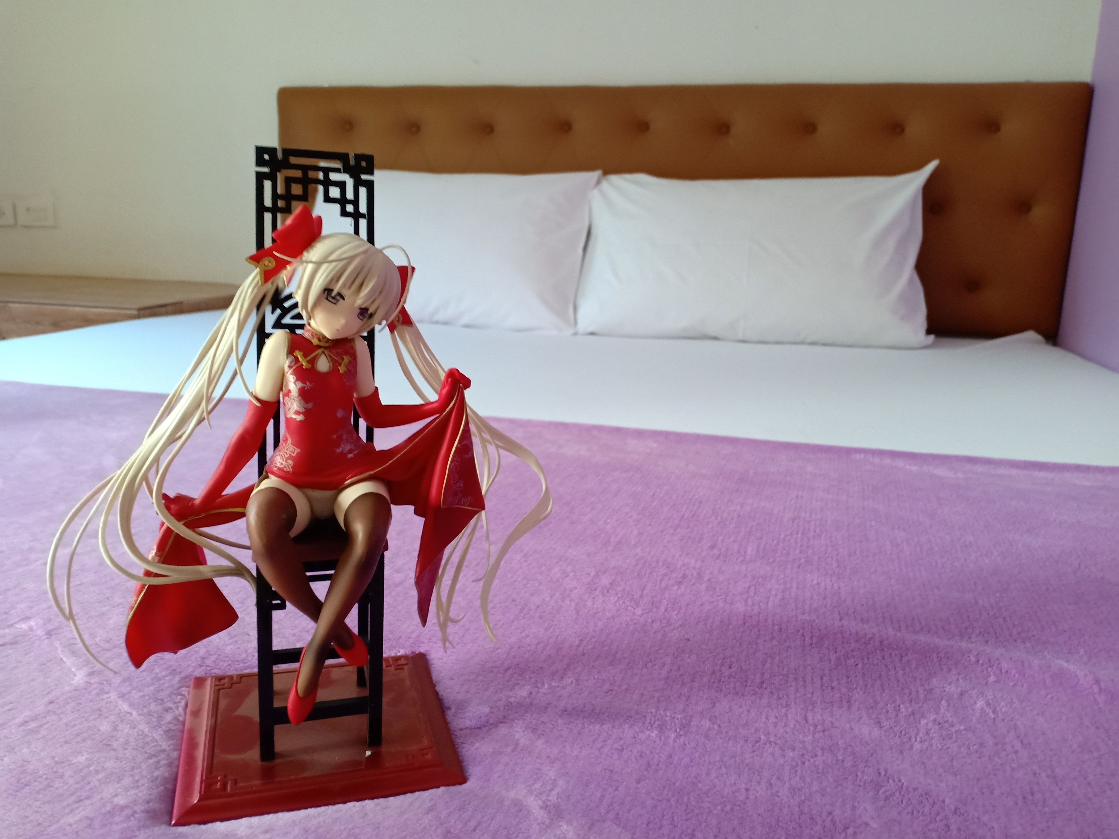 Un lit avec une tête de lit marron et des oreillers blancs, une couverture violette et une figurine posée sur un présentoir rouge.