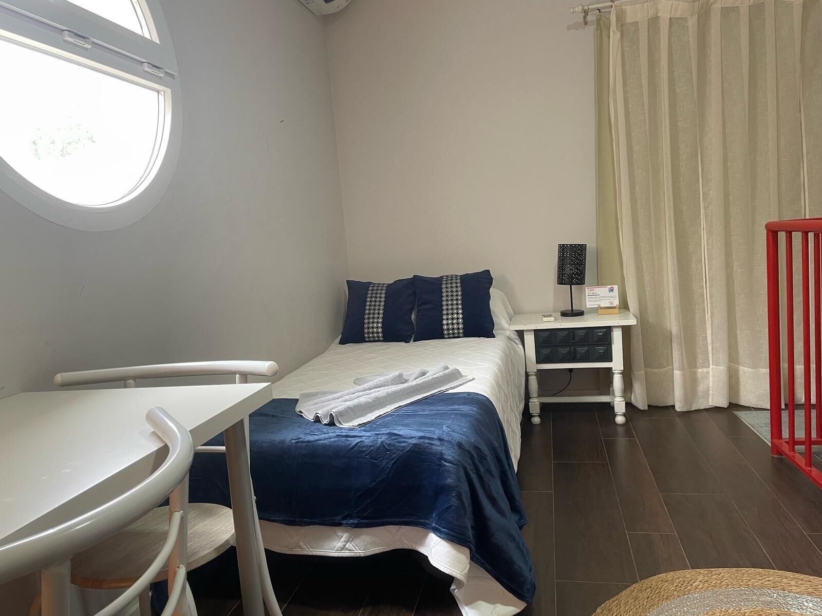 Una habitación pequeña y luminosa con una cama, un escritorio y una silla. Cuenta con una ventana que deja entrar luz natural, una cortina y una mesita de noche con una lámpara.