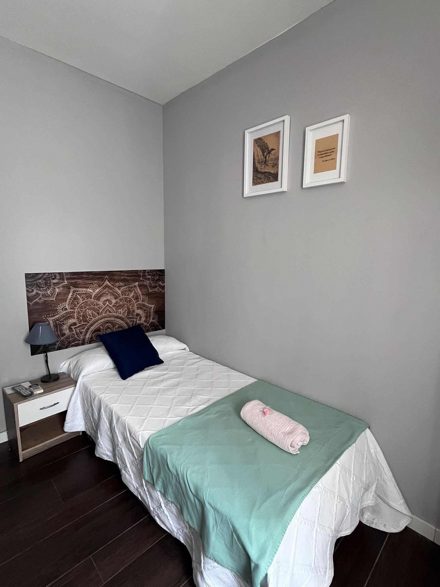 Un dormitorio ordenado con una cama, una mesita de noche, una lámpara y dos cuadros en la pared.