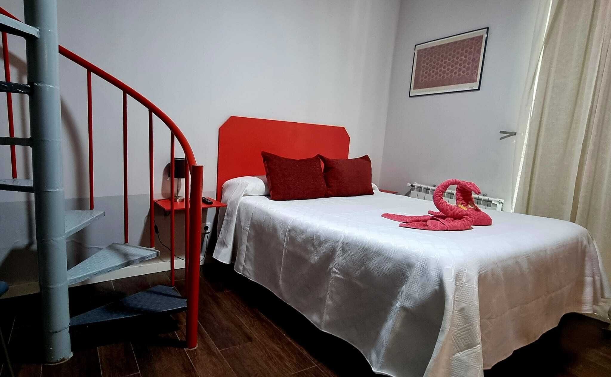 Habitación de hotel con cama, cabecera roja, escalera espiral roja, una silla roja, una toalla roja y un cuadro enmarcado en la pared.