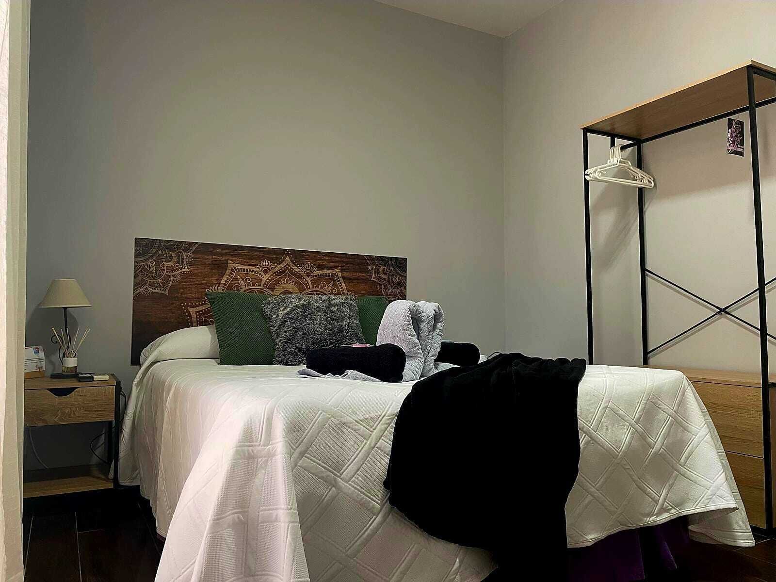 Un lit bien fait, avec une tête de lit à motifs, une table de chevet avec une lampe et un portant à vêtements suspendu.