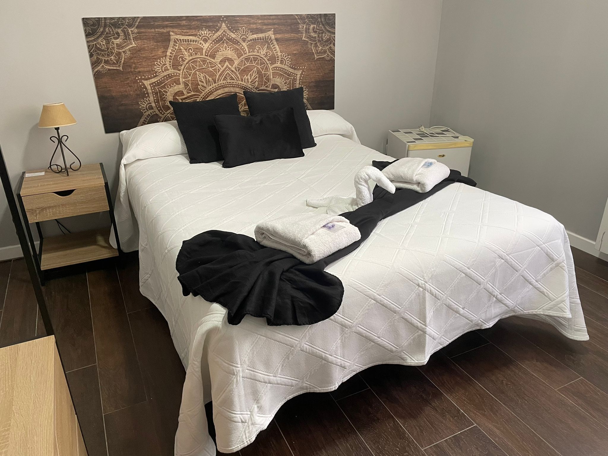 A neatly made bed with white bedding and black pillows, a bedside table with a lamp, and a wall tapestry.