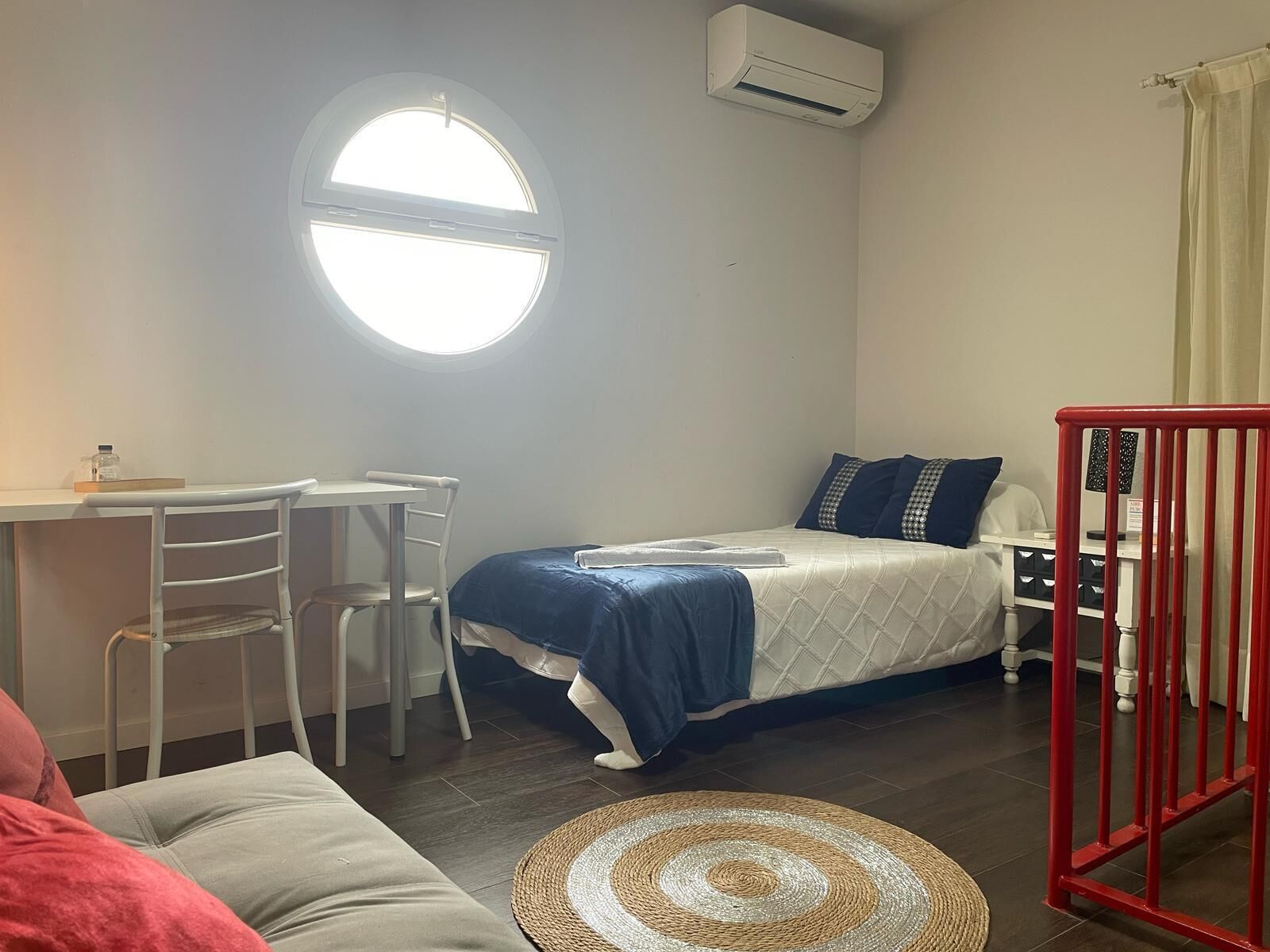 Una habitación pequeña y luminosa con un ventanal redondo, una cama con ropa de cama azul, un escritorio con una silla y una barandilla roja.