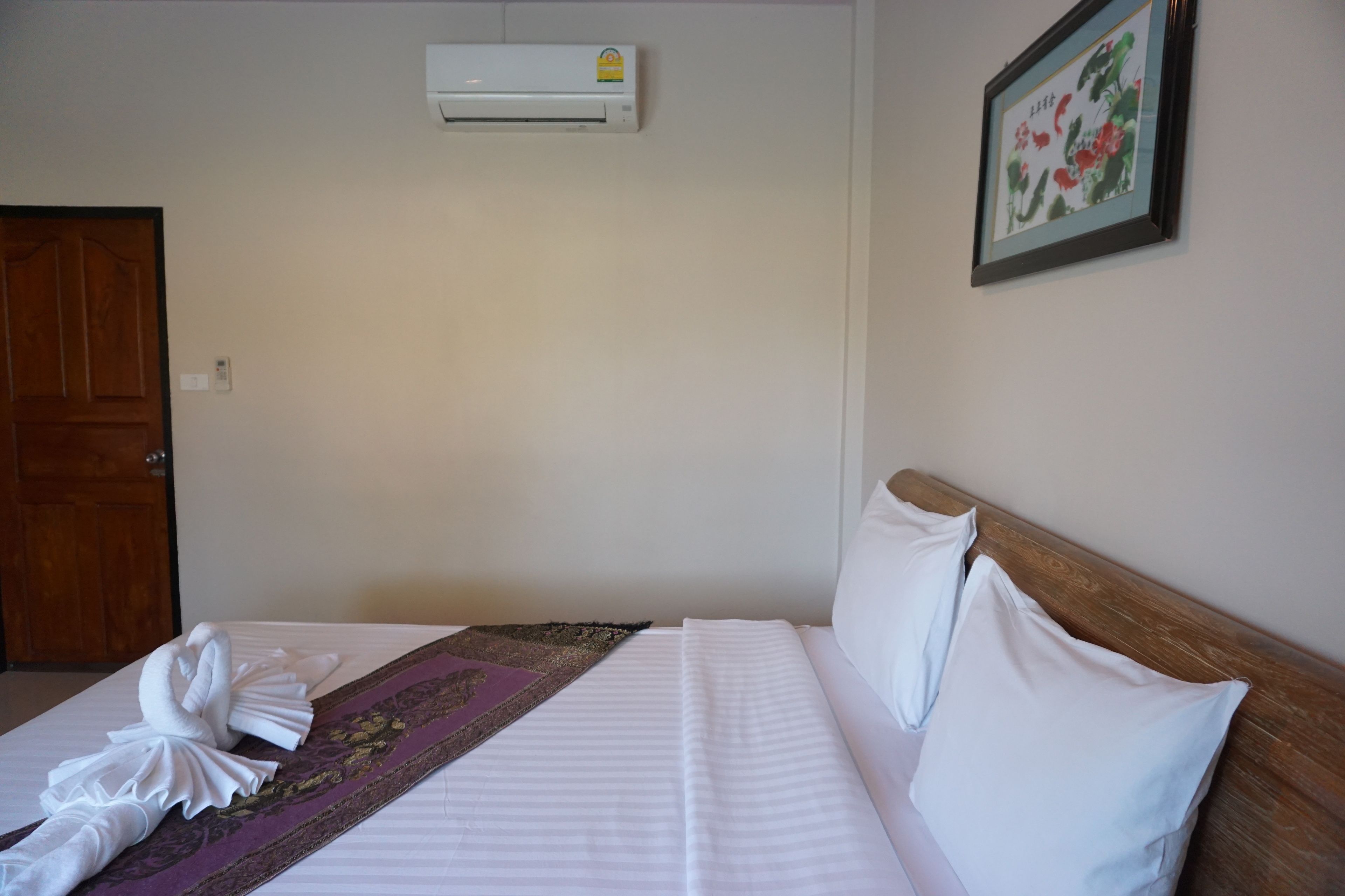 A hotel room with a bed, a wooden headboard, a wall-mounted air conditioner, a framed picture, and a door.