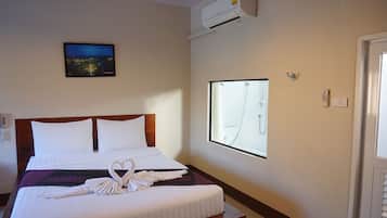 A hotel room with a bed, a window with a shower, an air conditioner, and a painting on the wall.