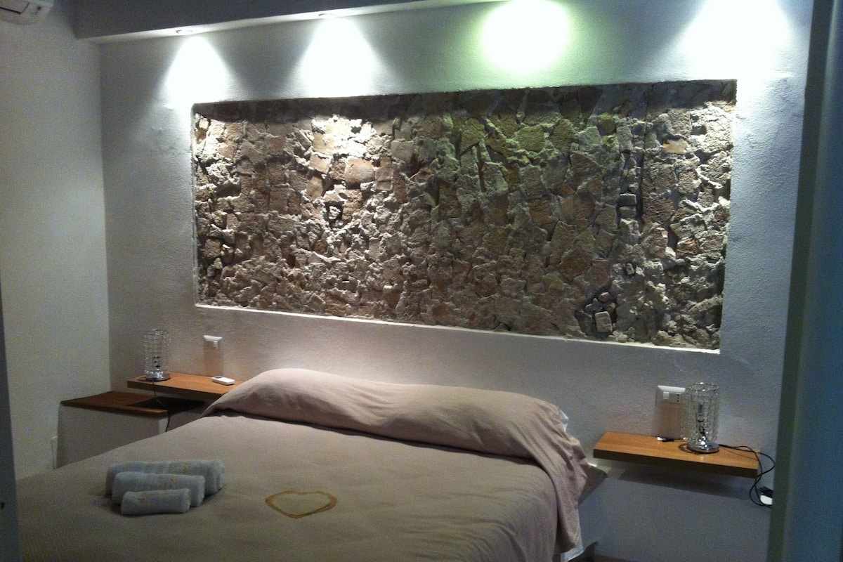 Une chambre à coucher avec une tête de lit en pierre, un lit agrémenté d’un coussin en forme de cœur et des tables de chevet avec des lampes.