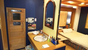 A small, compact room with a wooden door labeled "SAUNA," a sink with toiletries, a mirror, and a bed with a patterned bedspread. - HOTEL ESSOR - Adult Only (Nara)