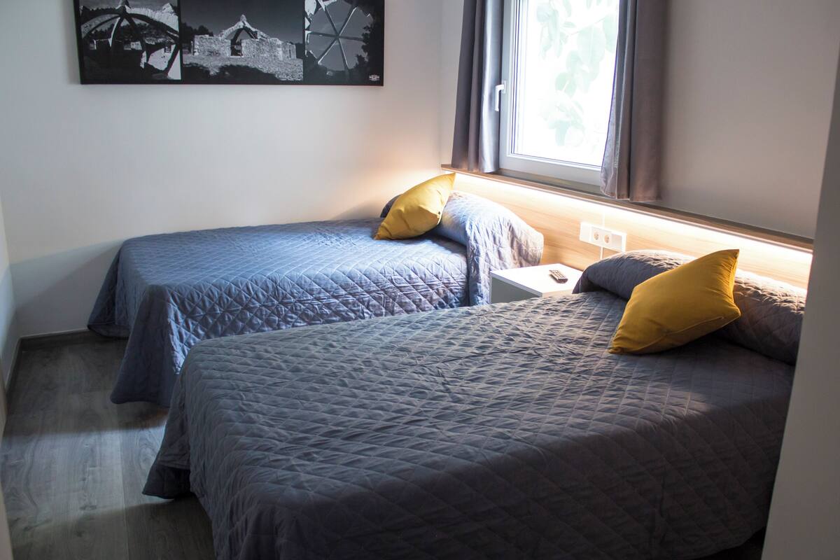 Habitación con dos camas, una ventana con cortinas, un enchufe en la pared y una obra de arte blanco y negro.