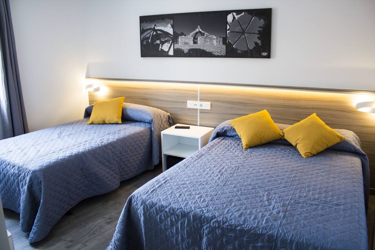 Habitación de hotel con dos camas, cada una con colcha azul acolchada y almohada amarilla. Hay una mesita de noche blanca entre las camas, y una obra de arte colgada en la pared sobre ellas.