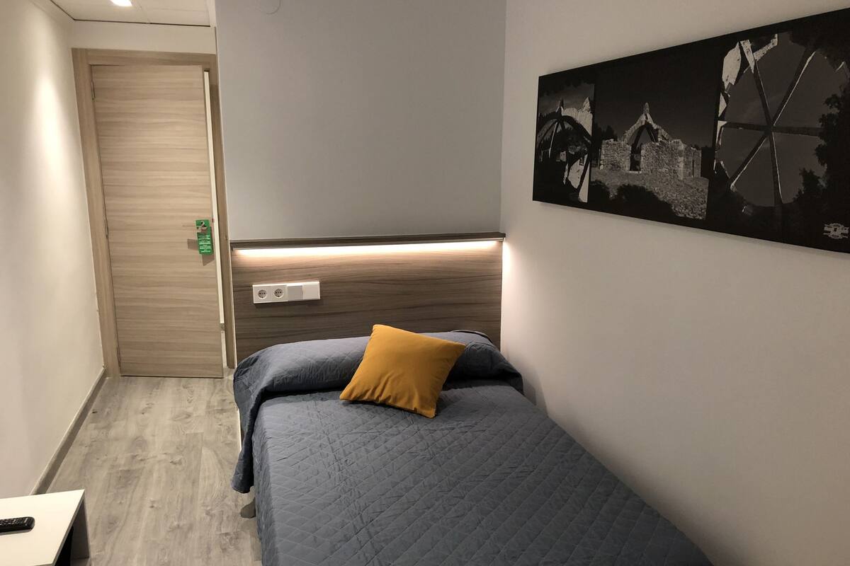 Habitación de hotel con cama, mesita de noche, lámpara de pared, un cuadro y una puerta.