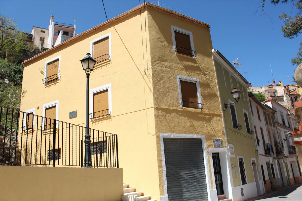 Un edificio amarillo con varias ventanas y balcones, una puerta con persiana cerrada y una cerca de metal.