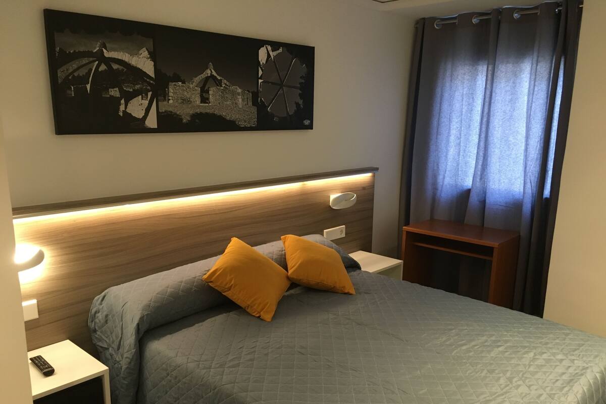 Una cama bien hecha con una colcha gris acolchada y dos almohadas naranjas, una mesita de noche con un control remoto, una lámpara de pared y un cuadro sobre la cama.