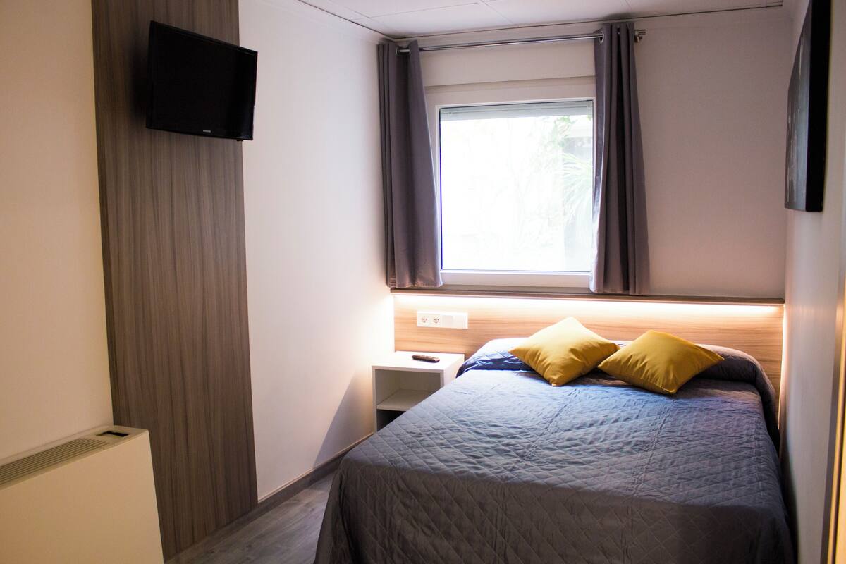 Un dormitorio con cama, mesita de noche, televisor montado en la pared y ventana con cortinas.