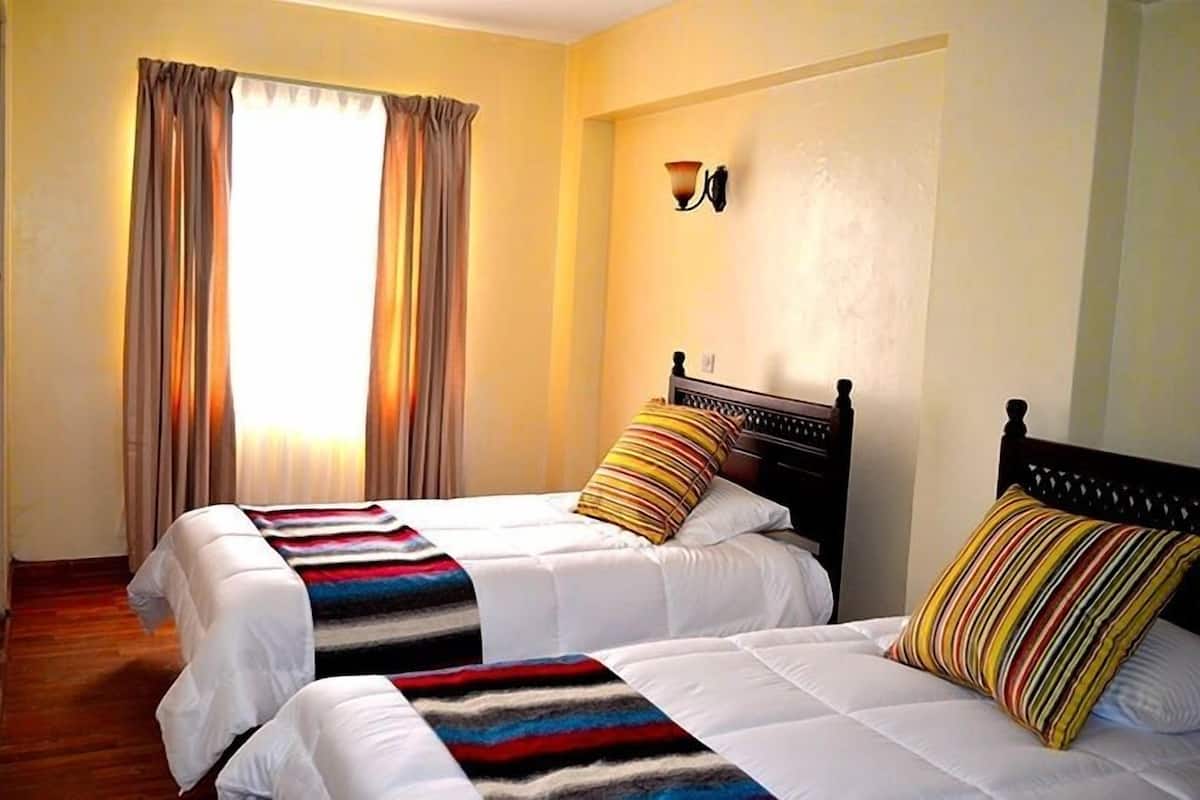 A hotel room with two beds, a large window with curtains, a wall-mounted light fixture, and a wooden headboard.