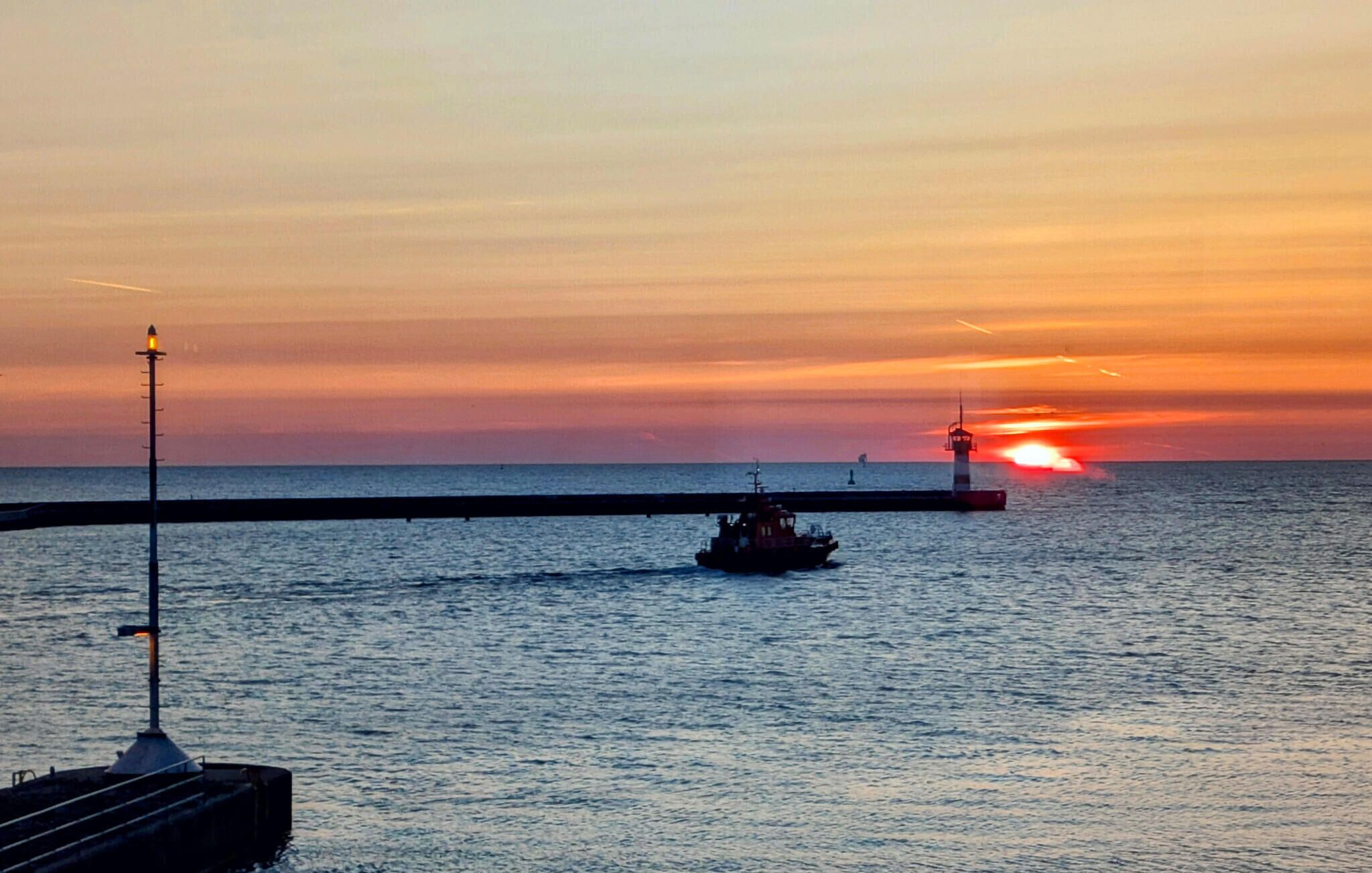A pier extending into calm waters at sunset with a boat and a lighthouse in the distance.