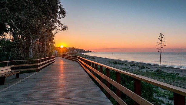 Una pasarela de madera que lleva a una playa con el atardecer de fondo.