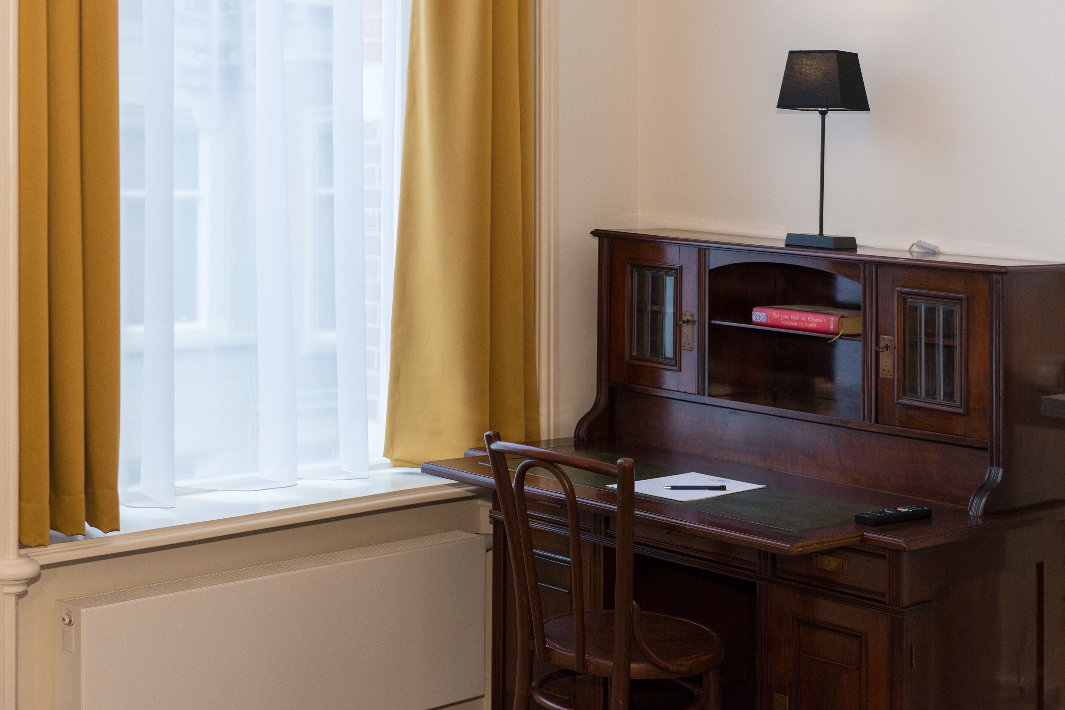 Een kamer met een houten bureau, een stoel, een lamp en een raam met gele gordijnen.