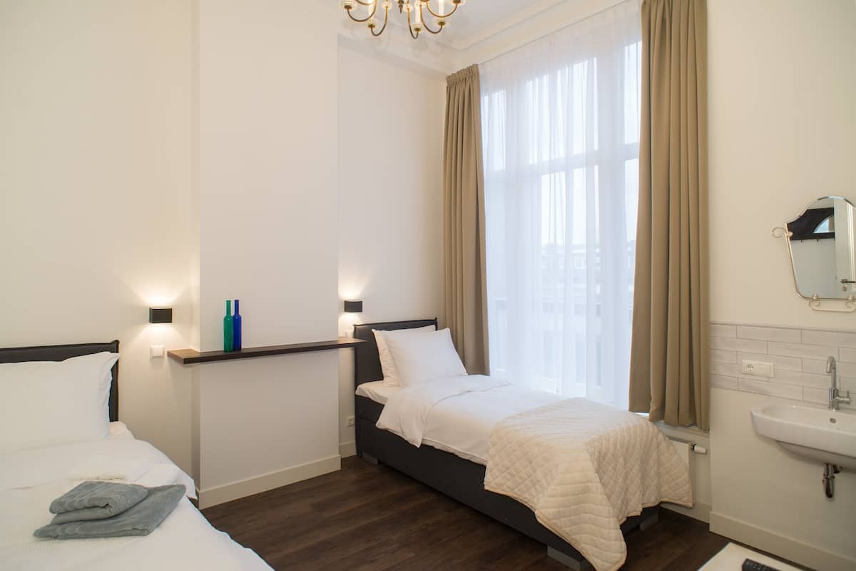 Une chambre d’hôtel comprenant un lit, une table de chevet, une lampe, une fenêtre avec des rideaux et une salle de bain avec un lavabo et un miroir.
