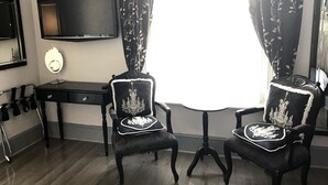 A room with two upholstered chairs with decorative trim, a small table with a lamp, a flat-screen TV mounted on the wall, and a window with patterned curtains. - Canterbury Lodge (Canterbury)